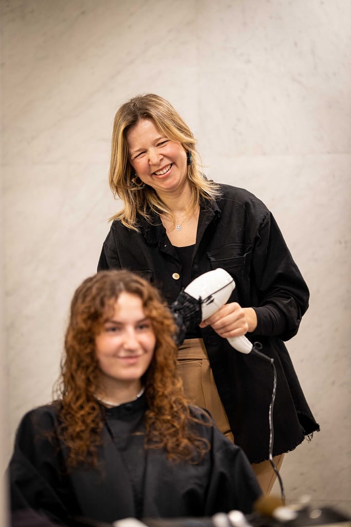 Coiffeuse à Paris 6 séchant les cheveux avec un séchoir blanc et noire. Les 2 femmes sont souriantes et contente de la coupe de cheveux qui vient d'être faites sur une chevelure ondulées et rousse.