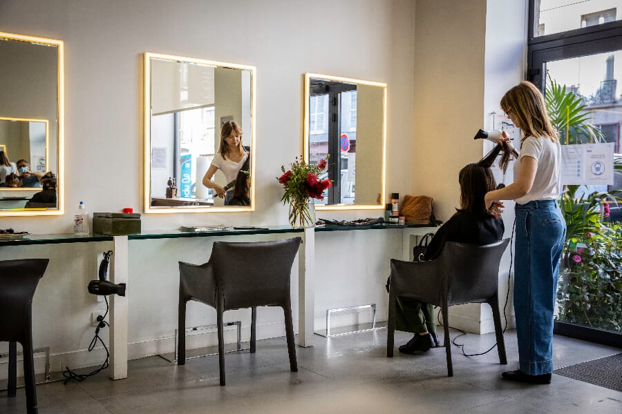 Meilleur Coiffeur Paris 5 coiffant face à des miroirs doré dans une ambiance lumineuse. Ils sont entouré de fleurs verte et violette.